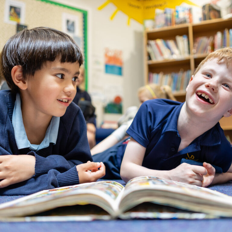 Two young boys lie on a blue carpet, reading a large open book together in a cozy library classroom, smiling.